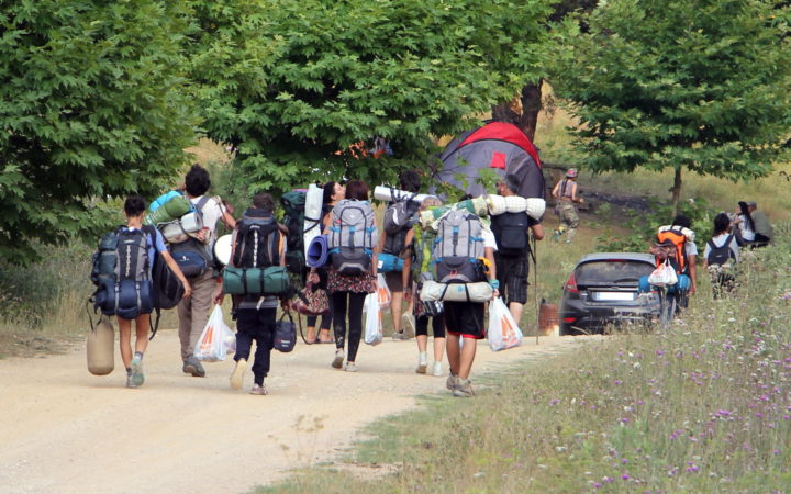Menschen mit Rucksäcken, Zelten und Schlafsäcken laufen einen Weg entlang